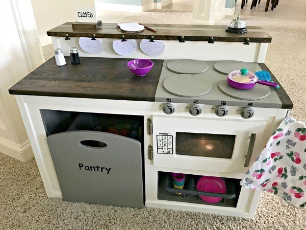 A super cute white kids play kitchen DIY with Farmhouse Style and restaurant menu board. I built this for about $50 and it's so cute. Kids love the food pantry, diner menu, dinner bell, motion-activated microwave light, open and closed sign.